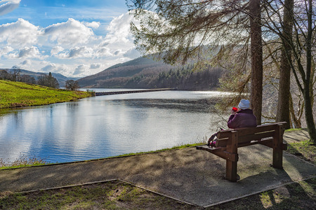 Landscape with a Lady relaxing in the cold weather sat on a bench overlooking a lake or reservoir drinking from a red cupの写真素材