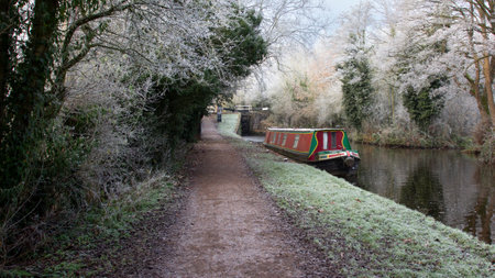 Looking down a canal from the towpath. It is taken on a frosty day and there is ice on the water and frost on the trees. A red narrow boat is moored.の写真素材