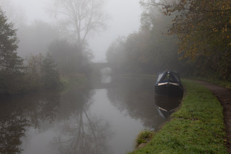 An atmospheric landscape looking down a canal. The image  was taken on a foggy morning, there is a narrow boat moored  and a bridge in the distanceの写真素材