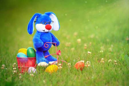 Blue easter bunny wearing glasses riding a tricycle with a basket full of colorful easter eggs in...