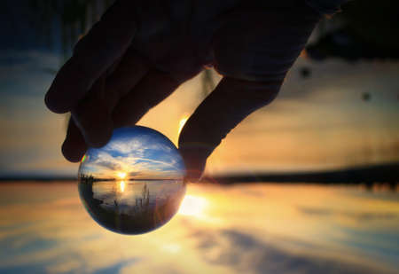 Fingers holding transparent glass ball reflecting the sunset on the water of a lakeの写真素材