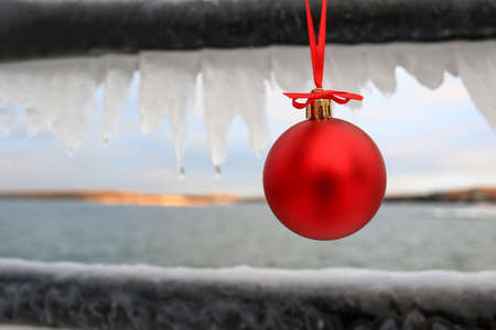 Red Christmas ornament hanging on an ice covered rail in front of a winter lake sceneの写真素材