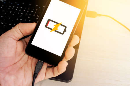man's hand holding smart phone is charging from laptop sunlight color