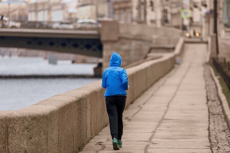 The girl is engaged in sports, jogging on the embankment. A woman in a blue jacket runs around the city.の写真素材