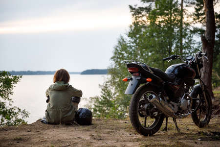 The girl biker sits with her back to the camera and looks at the lake. parked motorcycle in nature.の写真素材