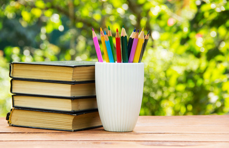 A stack of books and stack of colored pencils on natural blur green background. White porcelain cup with ribbed surface. Stationery on a wooden table. Garden in the background. Copy space.の写真素材