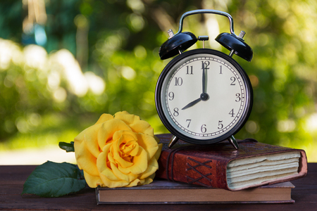 An old alarm clock, a stack of books and a rose on a wooden table. Books, a watch and a rose on a...