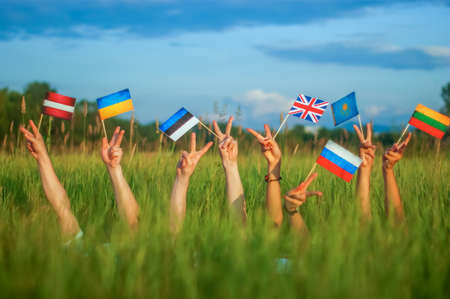 A group of young people holding different country flags and making peace or victory gesture on green...