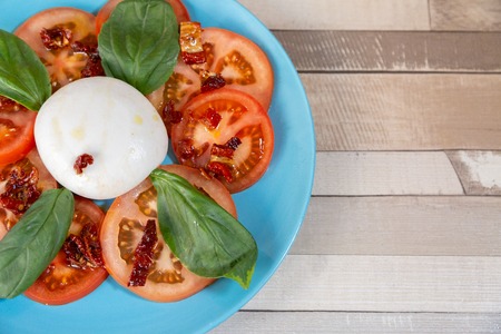 Close up of a half blue plate with fresh tomato, basil leaves, dry tomato and burrata cheese. Italian food in a wooden table. With copy spaceの写真素材