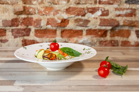 A white plate with rice and fresh vegetables. Vegan fresh food. Garnish with oregano and cherry tomatoes. In a wooden table and a brick wallの写真素材