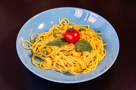 Yellow spaghetti with prawns, a cherry tomato and basil in a blue plate on a black table.の写真素材