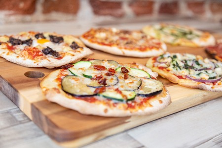 five pizzas on a wooden board with focus in zucchini and eggplant pizza in a wooden table with a brick wallの写真素材