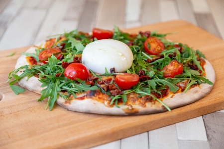a pizza on a wooden board with cherry tomatoes, arugula, and burrata cheese on a wooden table