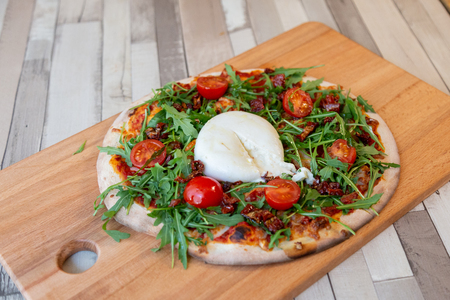 A pizza on a wooden board with cherry tomatoes, arugula, and burrata creamy cheese on a wooden table and brick wall for backgroundの写真素材