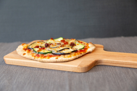 Side view of a pizza with zucchini, eggplant and dry tomatoes with gratin mozzarella cheese. On a wooden board and gray tableclothの写真素材