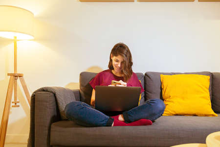Woman smiling on sofa looking at cell phone with laptopの写真素材