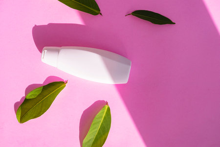 White cosmetic bottle container with green leaves on pink paper background. Natural organic beauty...