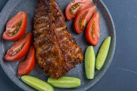baked pork ribs with honey lie with fresh sliced ââtomatoes and cucumbers on a dark gray concrete tabletopの写真素材
