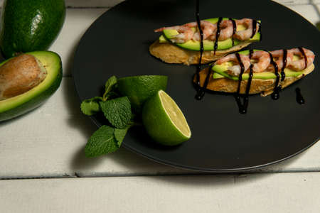 bruschetta with shrimps, avocado and lime on a dark plate on a white wooden backgroundの写真素材