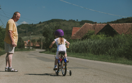 Little blonde girl riding a bicycle watched carefully by her grandfather, senior man, precious family time spent together, vintage color tonesの写真素材