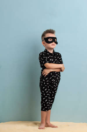 little boy in a black suit and a mask standing on sand on a light blue backgroundの写真素材