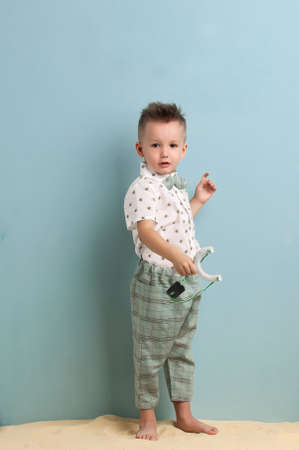 little boy in fashionable clothing and a slingshot in his hands stands on the sand on a light blue backgroundの写真素材