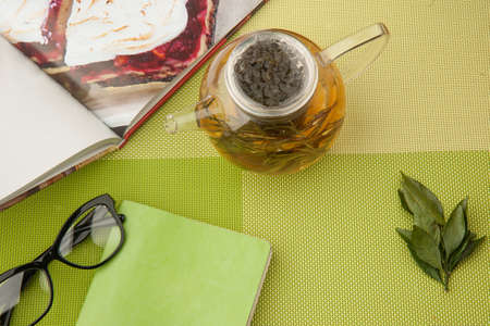 Transparent teapot with green tea with book and glasses on a green tableclothの写真素材
