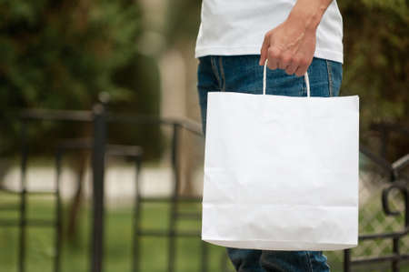 man with a white paper bag in hands on a park background. courier with a bag on the background of a green parkの写真素材