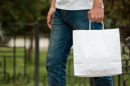 man with a white paper bag in hands on a park background. courier with a bag on the background of a green parkの写真素材