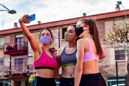 Three cheerful girls wearing masks take a selfie while practicing basketball on a court in the cityの写真素材