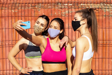 Three cheerful girls wearing masks take a selfie while practicing basketball on a court in the cityの写真素材