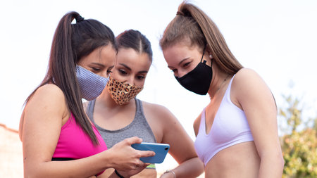 Three cheerful girls wearing masks look at a picture of a mobile phone while practicing basketball on a court in the cityの写真素材