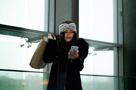 Latina girl is shopping in a mall, she looks at her cell phone and holds a shopping bag while smiling.の写真素材