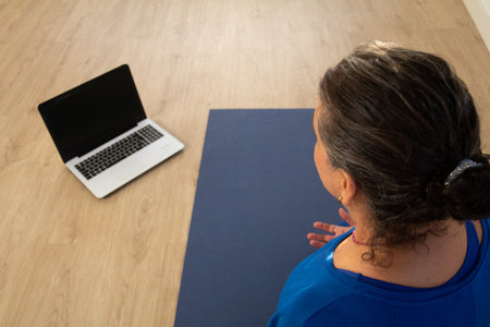 The yoga teacher, dressed in blue, is sitting on a mat looking at her computer to start the online class.の写真素材