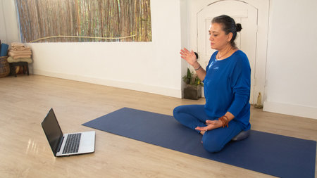 The Yoga teacher dressed in blue is sitting on a mat while chatting with her students on a laptop in a video call.の写真素材
