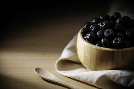 Fresh blueberries with small drops of water on the surface, in a bamboo bowl on a wooden table, with a tablecloth below and a wooden spoon.の写真素材