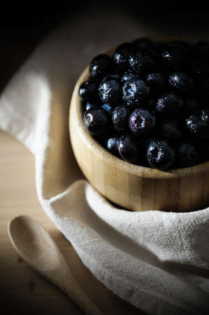 Fresh blueberries with small drops of water on the surface, in a bamboo bowl on a wooden table, with a tablecloth below and a wooden spoon.の写真素材