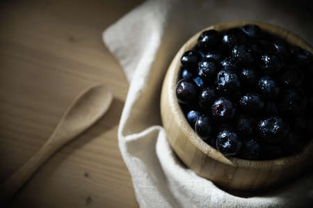 Fresh blueberries with small drops of water on the surface, in a bamboo bowl on a wooden table, with a tablecloth below and a wooden spoon.の写真素材