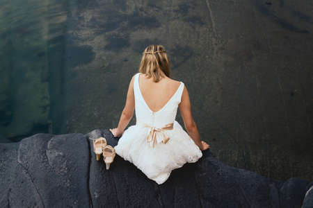 Woman wearing a white dress sitting on the rock looking at the waterの写真素材