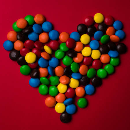 Frame of red, blue, orange, yellow, brown and green rounded candy on a red background with the shape of a heart.の写真素材