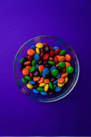 Glass bowl filled with blue, orange, yellow, red, brown and green candy on a purple background seen...