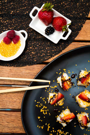 Sweet and delicious sushi with strawberries, blackberries, sesame seeds and chocolate dripping on a black plate and a black tablecloth with chopsticks on wooden table.の写真素材