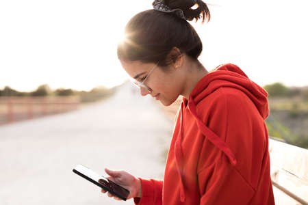 Young woman with glasses on her twenties chatting on her phone while sitting in a bench of a park. She is smiling and wearing a red sweater and a ponytail. She is backlit with daylight at sunsetの写真素材