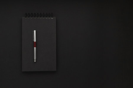 Black spiral notebook with a silver fancy pen on top of it in the middle of a black backdrop....