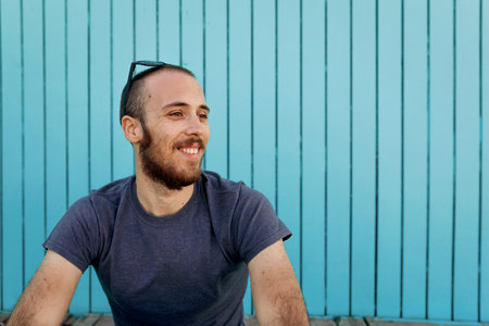 Happy young bearded with sunglasses over the head man is smiling at Isla Canela beach sitting in front of a wooden blue wall while looking awayの写真素材