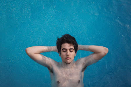 Top view of a young man floating in a swimming pool with his hands in the back of his head and his eyes open. Enjoying vacations while sunbathing in a hotel. Holiday concept.の写真素材