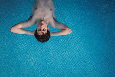 Top view of a young man floating in a swimming pool with his hands in the back of his head and his eyes open. Enjoying vacations while sunbathing in a hotel. Holiday concept.の写真素材