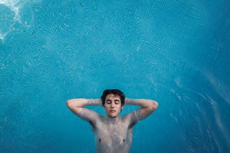 Top view of a young man floating in a swimming pool with his hands in the back of his head and his eyes open. Enjoying vacations while sunbathing in a hotel. Holiday concept.の写真素材
