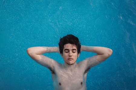 Top view of a young man floating in a swimming pool with his hands in the back of his head and his eyes open. Enjoying vacations while sunbathing in a hotel. Holiday concept.の写真素材