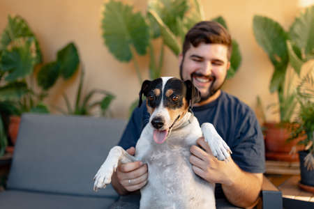 Young man sitting and smiling is holding up his dog while playing with his paws. The dog is looking...